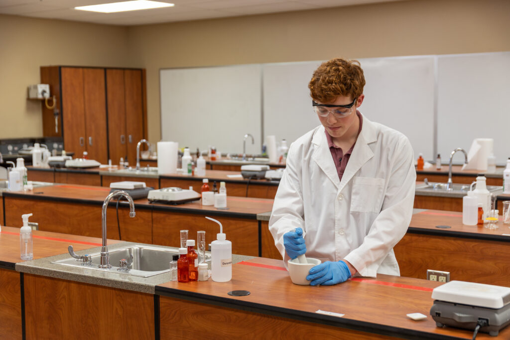 A person in a lab coat, safety goggles, and blue gloves uses a mortar and pestle in a spacious science laboratory—perfect for students preparing for the Pharmacy Early Admissions Program, with wooden cabinets, sinks, and bottles of chemicals on the benches.
