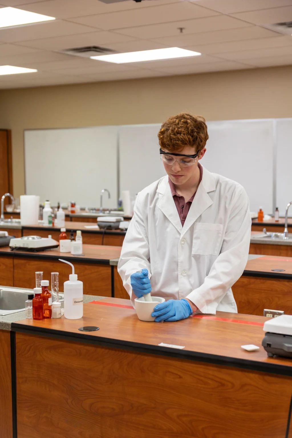 A person wearing a lab coat, gloves, and safety goggles uses a mortar and pestle at a lab bench in a science classroom. Various bottles and lab equipment are on the counter.