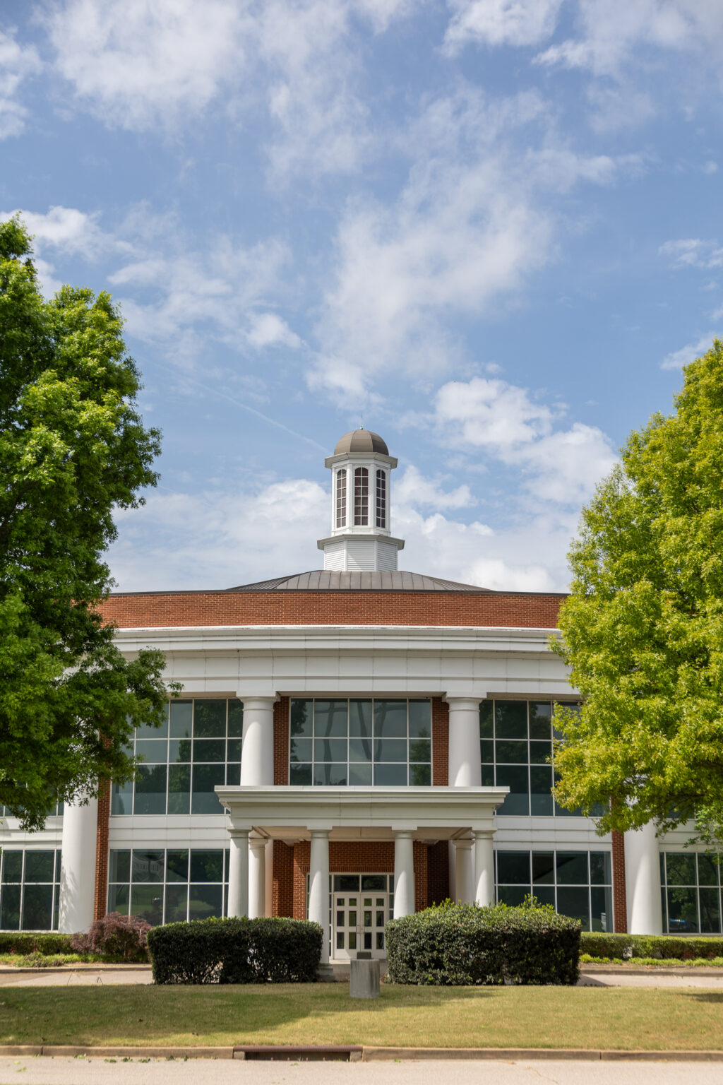 A brick and glass building on the Memphis Campus with a central entrance, large white columns, and a small cupola on the roof, framed by two green trees under a partly cloudy sky.