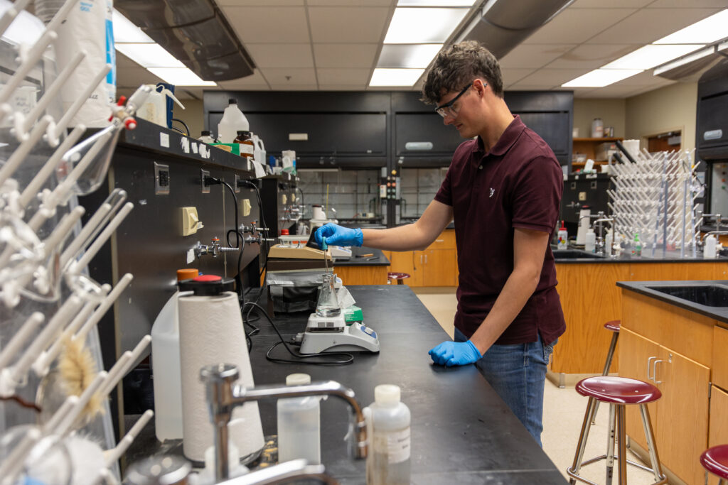 A person wearing blue gloves and a maroon shirt operates scientific equipment on a lab bench, conducting forensic analysis in a laboratory filled with glassware, bottles, and instruments.