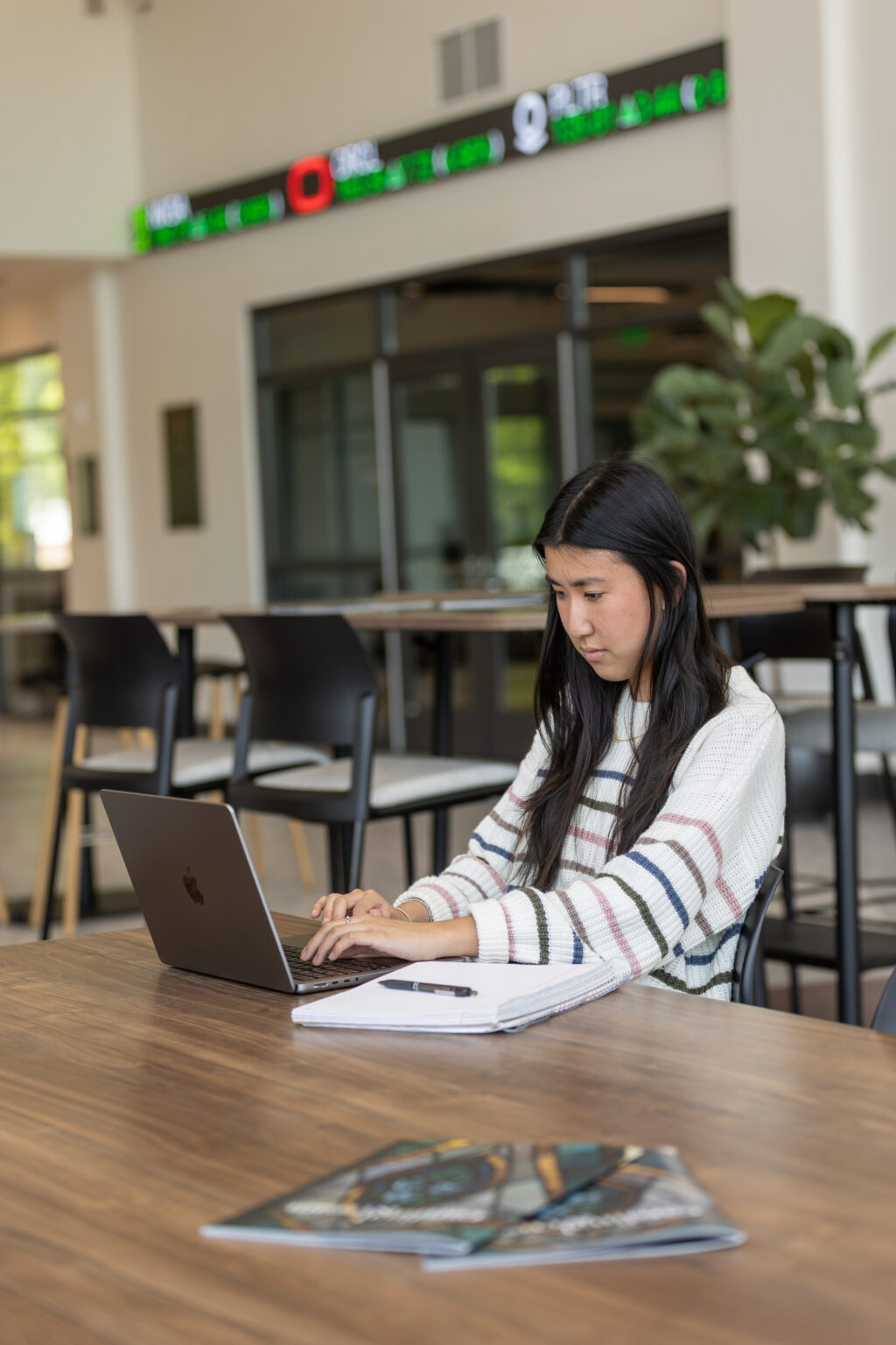 A woman with long dark hair sits at a wooden table working on a laptop. Surrounded by papers and magazines, she focuses on Business Administration as a digital stock ticker display glows in the background.