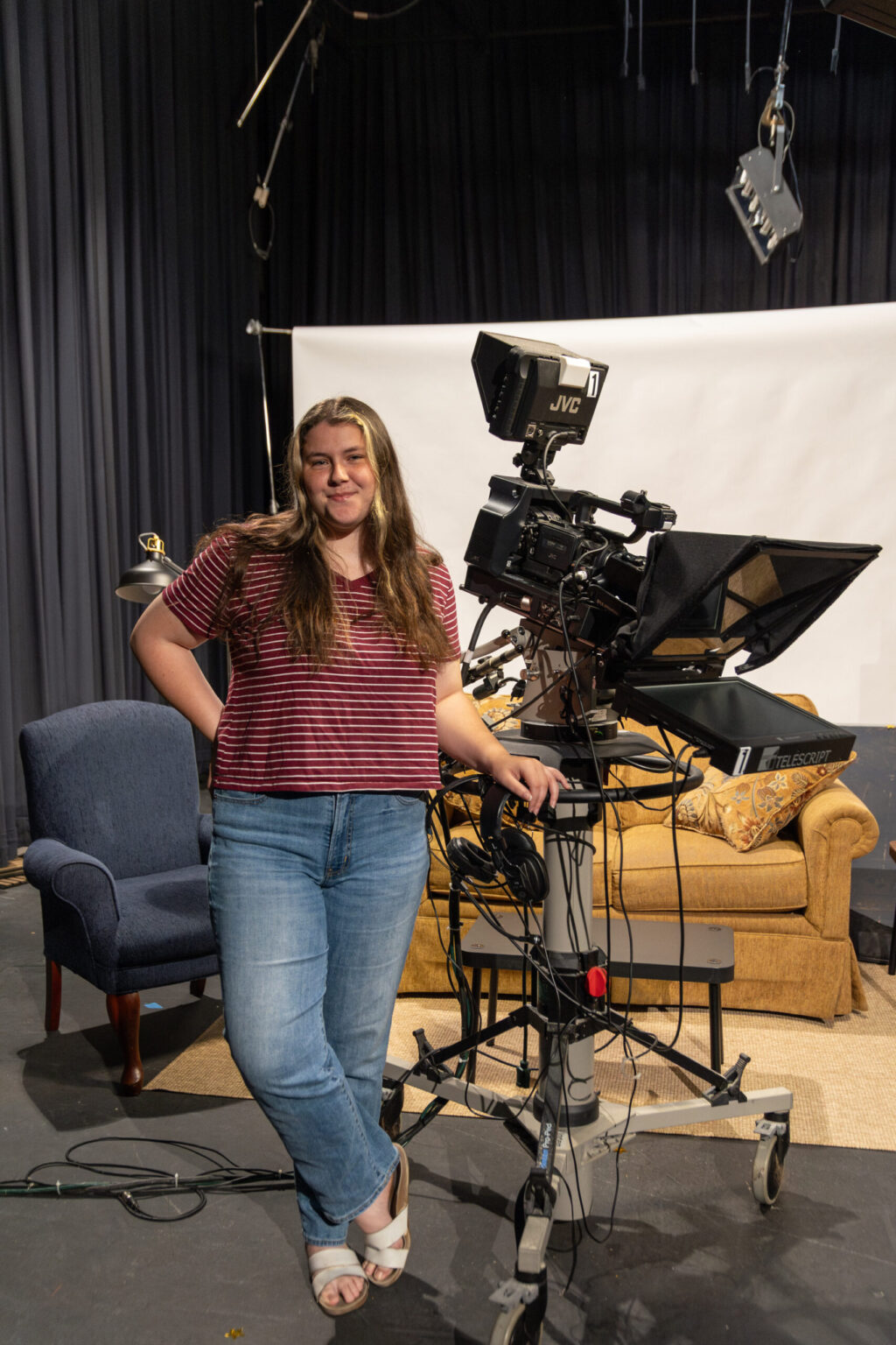 A person in a striped shirt and jeans stands next to a professional video camera on a set—perfect for Digital Media Studies—with a blue chair, tan couch, white backdrop, and visible studio lights and cables in the background.