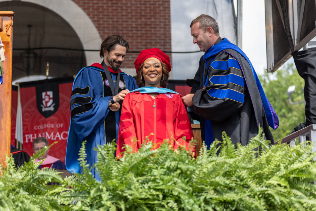 A woman earning her Doctor of Education (EdD) stands smiling between two people in blue and black robes at an outdoor graduation ceremony. One person places a hood over her shoulders, with a College of Pharmacy banner visible in the background.