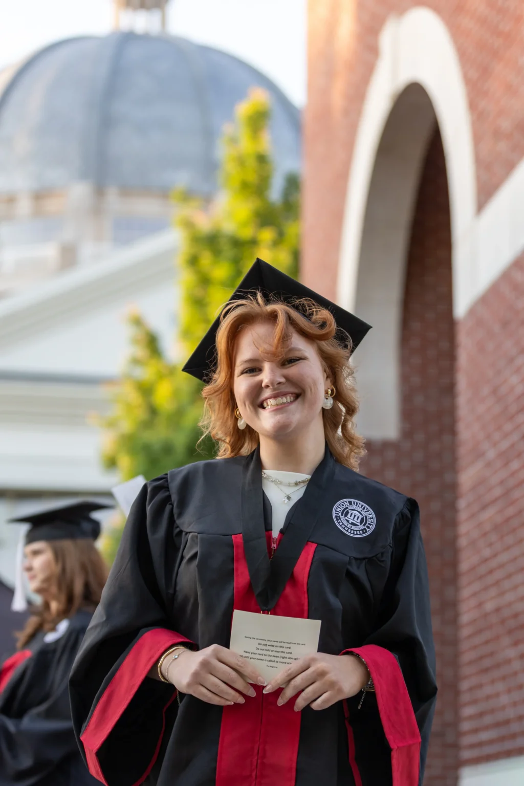 A graduate in a black cap and gown with red accents stands outside, smiling at the camera and holding a document. A large building with a dome and brick archway is visible in the background.