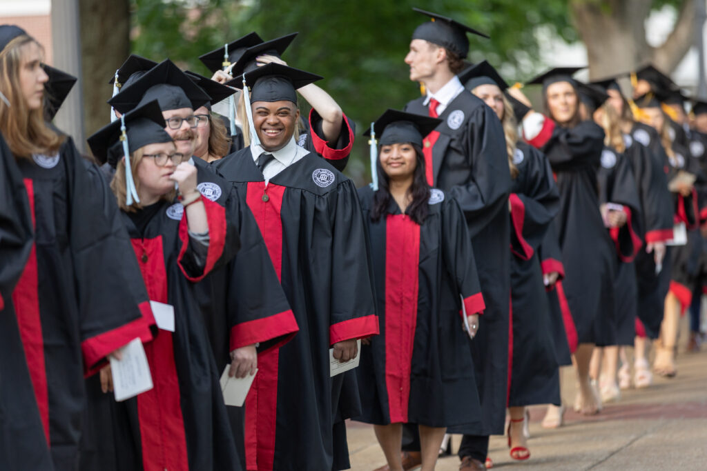 A group of graduates in black caps and gowns with red accents stand in a line outdoors, some smiling and looking at the camera, during a graduation ceremony.