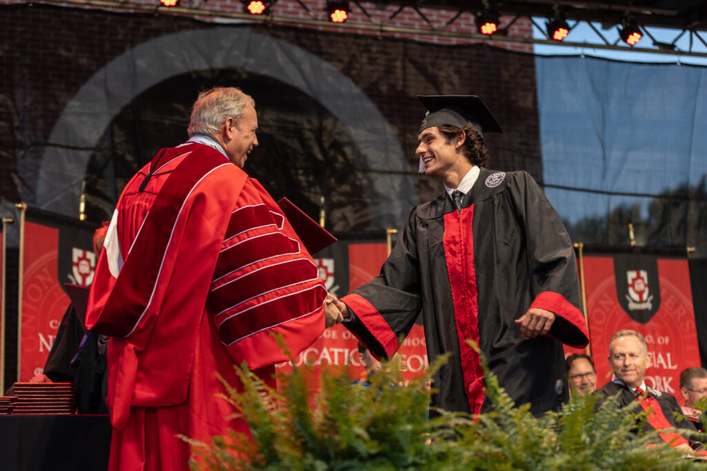 A university official in red academic regalia shakes hands with a graduating student in a black cap and gown on stage during a commencement ceremony. Other officials are seated in the background.