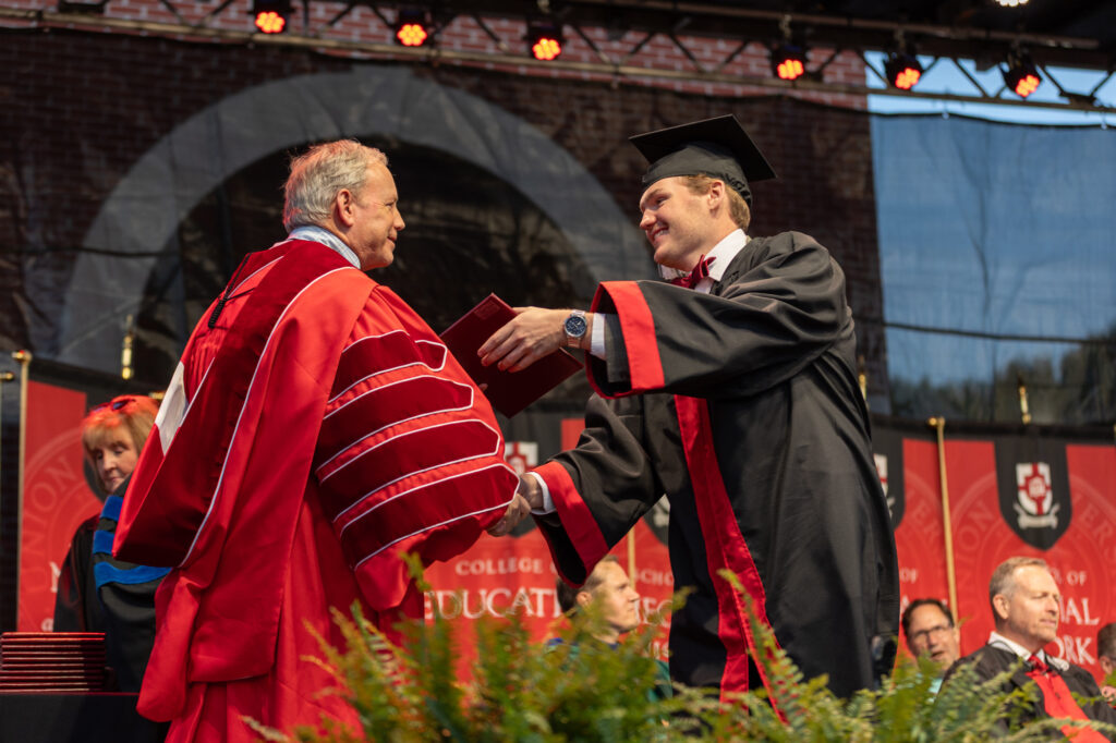 A man in a graduation cap and gown shakes hands with a university official in red academic regalia while receiving a diploma on stage at a commencement ceremony. Other officials and banners are visible in the background.