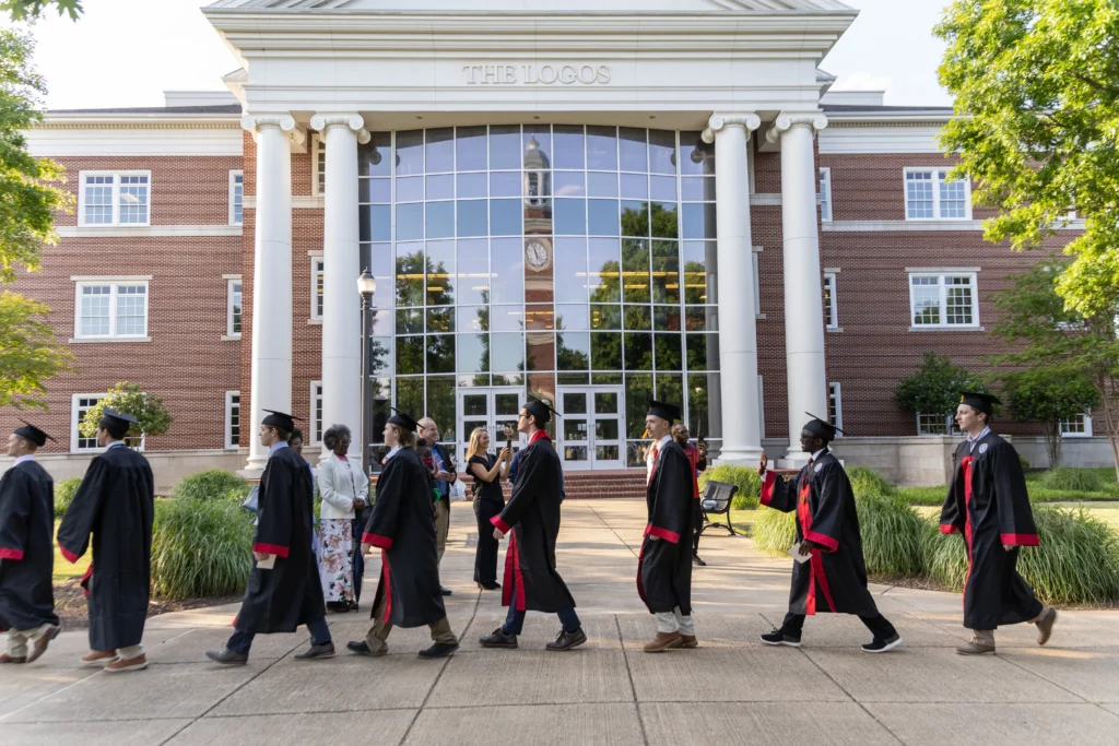 A group of graduates in caps and gowns walk in a line outside a brick academic building with white columns and large windows, while others stand nearby. It is sunny and trees frame the scene.