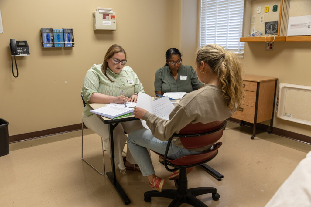 Two women sit at a desk reviewing papers with a third woman sitting across from them in a medical office. The room, which appears to be set up for consultations related to the Nursing Post-Graduate Certificate program, features medical equipment on the walls and documents on the table.