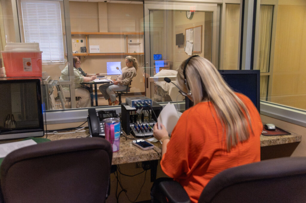 A woman in an orange shirt monitors and takes notes at a desk with equipment, observing two people having a conversation in a room through a large window—reflecting the careful supervision and analytical skills developed in a Nursing Post-Graduate Certificate program.