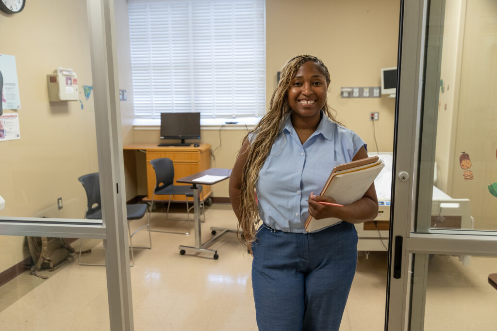 A woman stands inside an office or classroom, smiling and holding folders, possibly related to her Post-Graduate Certificate in Nursing. She is wearing a light blue blouse and blue pants. Desks, chairs, and office equipment fill the background.