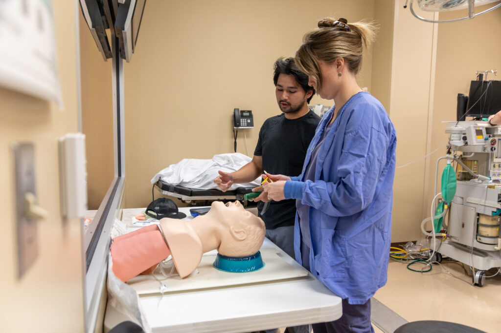 Two people stand by a medical training mannequin on a table in a clinical room, demonstrating key concepts in nurse anesthesia. One person, possibly a DNP student, holds medical equipment while the other observes closely. Medical supplies and equipment are visible in the background.