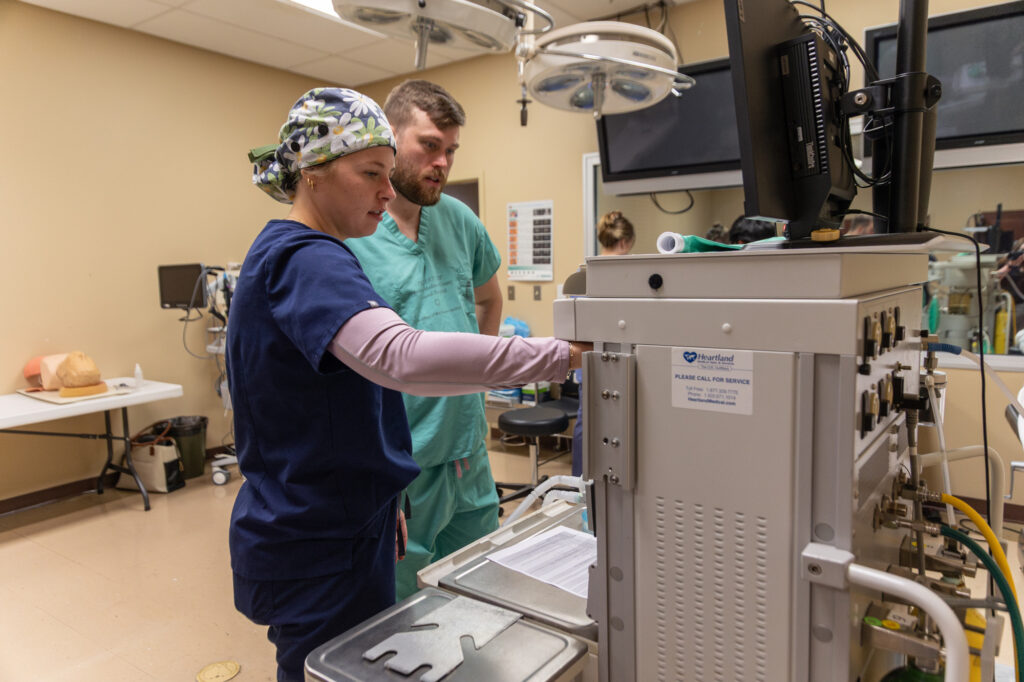 Two medical professionals in scrubs, including a DNP specializing in Nurse Anesthesia, examine and discuss a medical device in a clinical room. Both focus intently on the machine's controls while medical equipment, monitors, and supplies are visible in the background.
