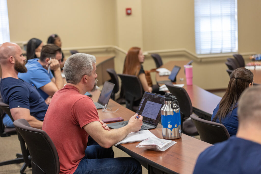 Several adults, including a Nurse Practitioner, sit at desks in a classroom with beige walls and large windows. Some use laptops and tablets while one writes in a notebook, creating a focused learning environment.