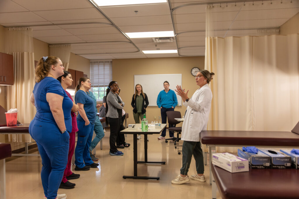 A Nurse Practitioner in a white coat speaks to a group of people in scrubs gathered in a medical training room. Tables, medical supplies, and a clock are visible in the background.