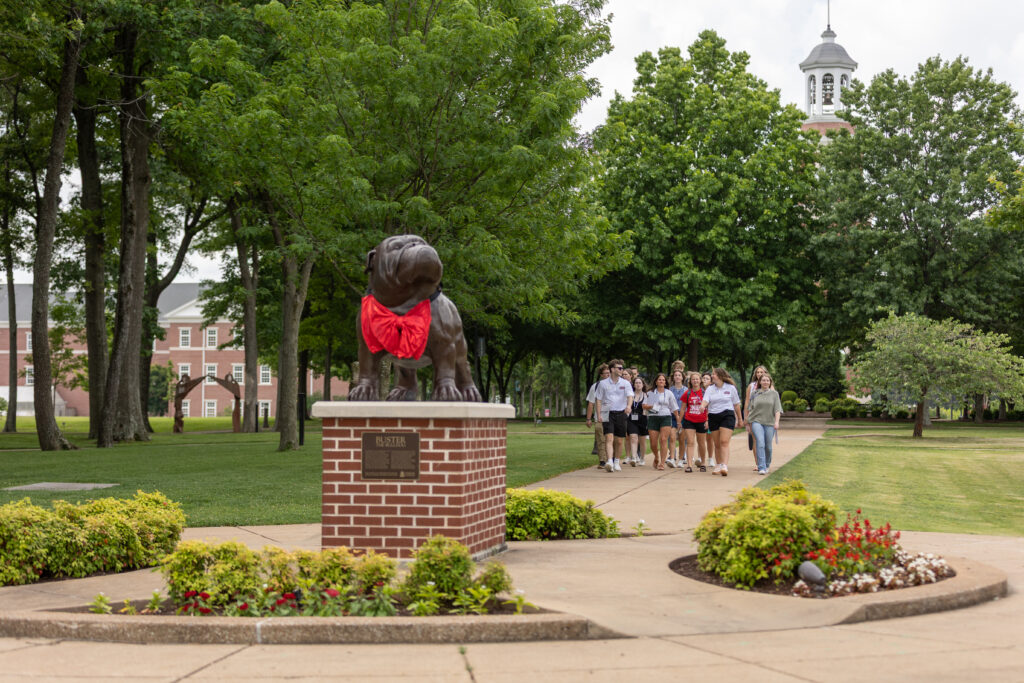 A group of people walk along a tree-lined path toward a statue of a bulldog wearing a red cloth around its neck on Union's main campus in Jackson, with buildings and a clock tower visible in the background.