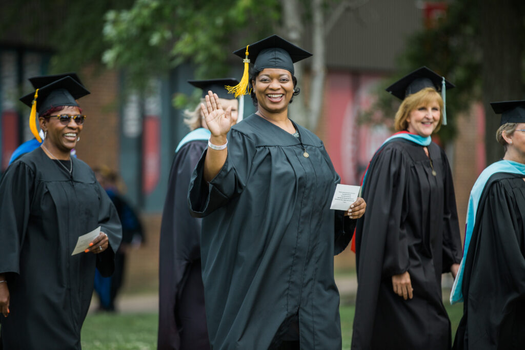 A group of adults in graduation gowns and caps walk outdoors, celebrating their achievements in adult education. One woman in the center smiles and waves at the camera, holding a piece of paper. Trees and buildings are visible in the background.