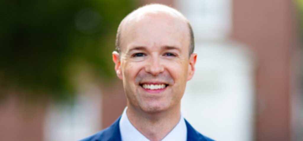 A man with a bald head and light skin is smiling, wearing a blue suit and white shirt. The blurred background with green and brown tones suggests an outdoor setting—perfect for a commencement ceremony celebrating Union graduates like Walker.