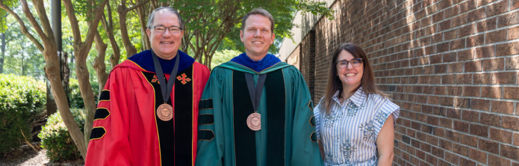 Three adults stand outdoors next to a brick wall and greenery. Two men wear academic robes and medals, while a woman in a striped dress stands beside them, all facing the camera and smiling.