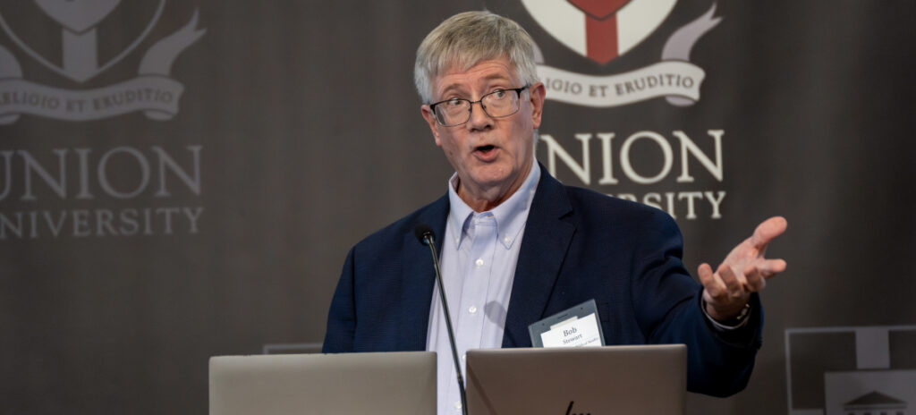 An older man, identified as Stewart, wearing glasses and a navy blazer speaks at a podium with microphones during a Bible Conference, gesturing with his right hand. The background displays the Union University logo.
