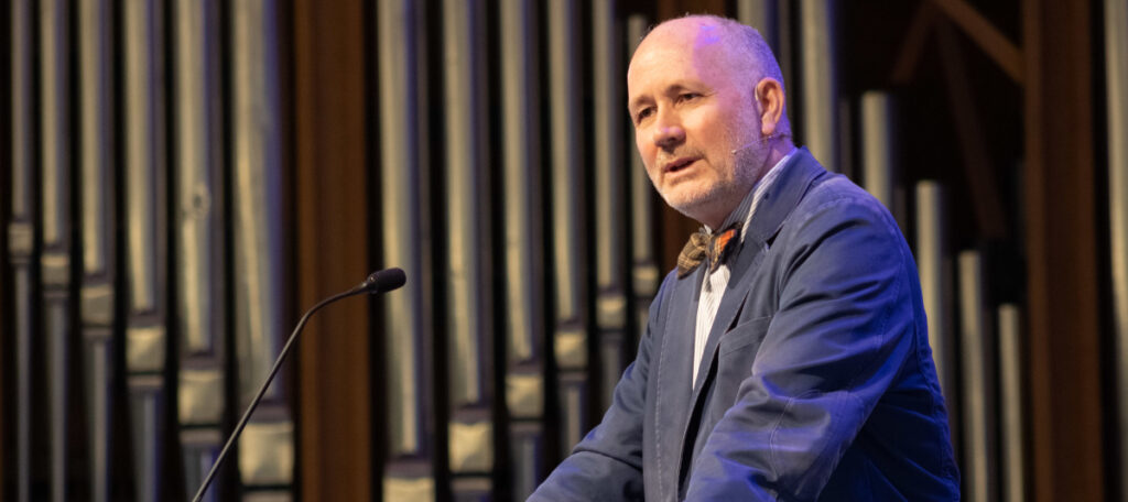 A man with a shaved head and grey beard, wearing a blue jacket, speaks at the podium during the Dockery Lectures. Large vertical pipes are visible in the background as he addresses the audience on March 19.