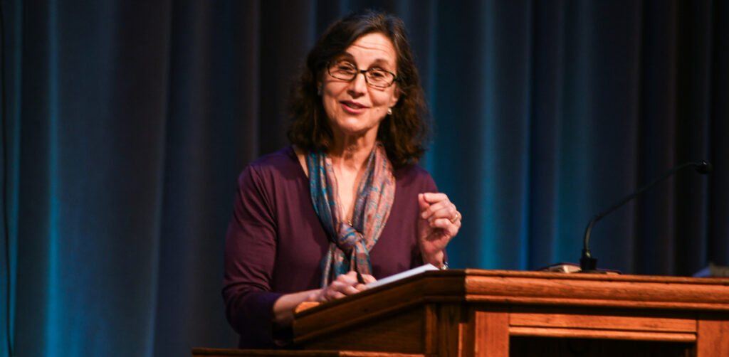 A woman with glasses and a scarf speaks at a wooden podium, addressing topics such as unbelief, while holding a pen and paper and wearing a microphone headset. Blue curtains are in the background.