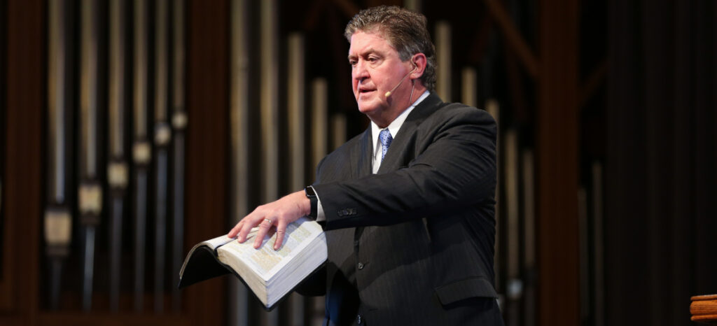 A man in a suit and tie, possibly Union alumnus Steve Gaines, stands speaking with an open book—likely a Bible—in one hand. He wears a headset microphone and is positioned in front of impressive organ pipes.