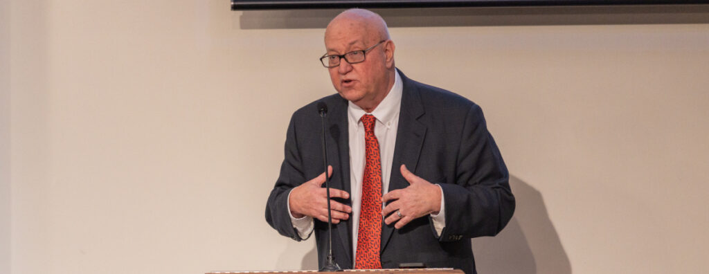 An older man in a dark suit, red patterned tie, and glasses speaks at a podium during a Union luncheon, gesturing with both hands. He stands in front of a light-colored wall.