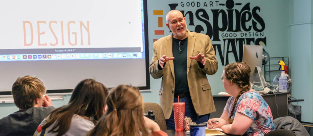 A man stands and speaks in front of a classroom with students seated at desks, facing him. The projector screen displays the word DESIGN. A wall behind him features inspirational words about Christ-centered excellence in art and design.
