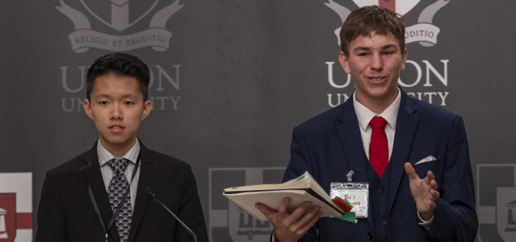 Two young men in suits stand at a podium in front of a Union University backdrop with a crest. One gestures while speaking during the debate tournament; the other, ready for NITOC, looks straight ahead holding a notebook.