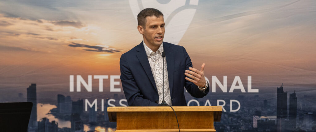 A man in a suit speaks at a podium with a microphone. Behind him, the cityscape backdrop and partial text INTERNATIONAL MISSION BOARD set the scene for the Powell Lecture on multicultural missions.