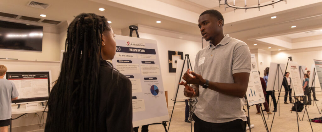 Two people stand in front of a presentation board discussing research at the Scholarship Symposium in a well-lit room, with other people and posters filling the Union in the background.