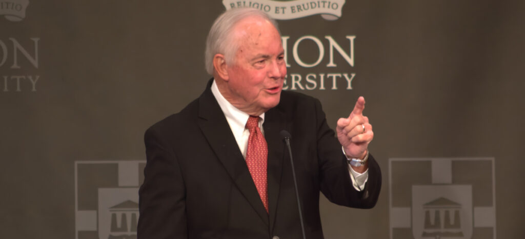 An older man in a black suit, white shirt, and red tie speaks at a podium during a Valentine's banquet, gesturing with his right hand. The background displays the words Union University and a university crest as West Tennessee pastors look on.