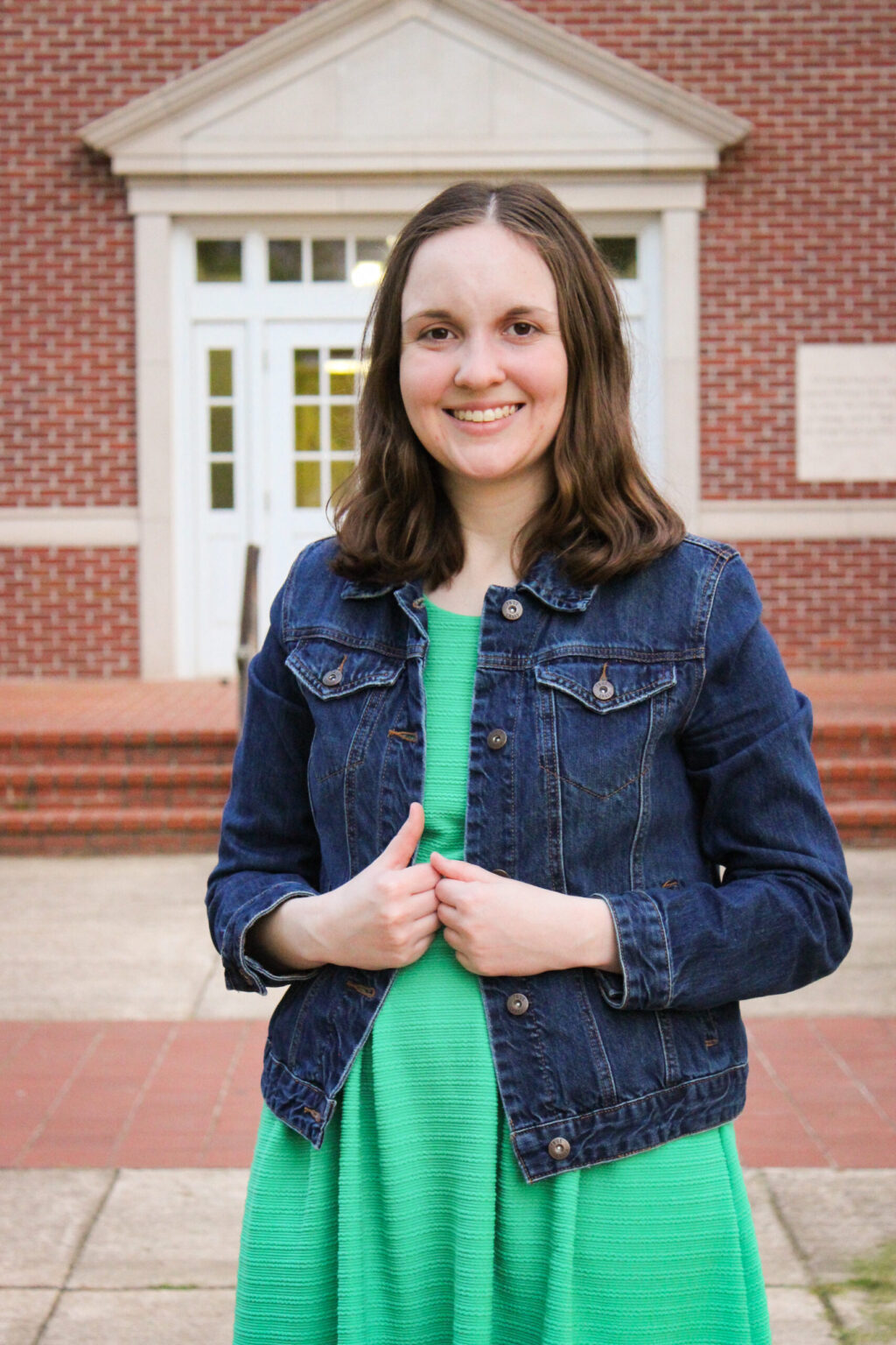 A woman with long brown hair, stands outdoors in front of a brick building with white-trimmed doors. She is wearing a green dress and a denim jacket, smiling warmly at the camera.