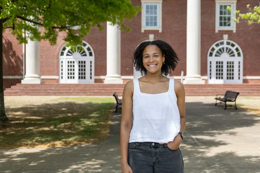 Aryn Roberts, with curly hair, wearing a white sleeveless top and dark jeans, stands smiling outside in front of a brick building with large windows and columns. Benches and trees are visible in the background.