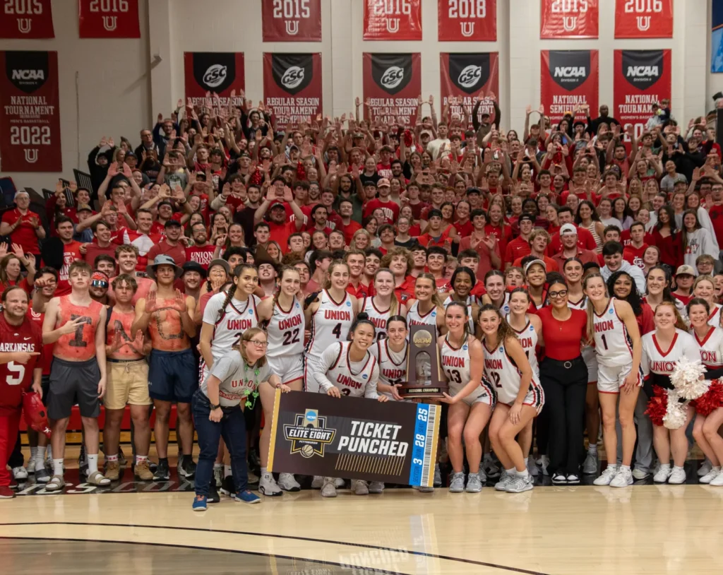 A large group of students, cheerleaders, and fans pose together on a gymnasium floor, many wearing red and white. Some hold a “Ticket Punched” sign. Red championship banners hang on the wall in the background.