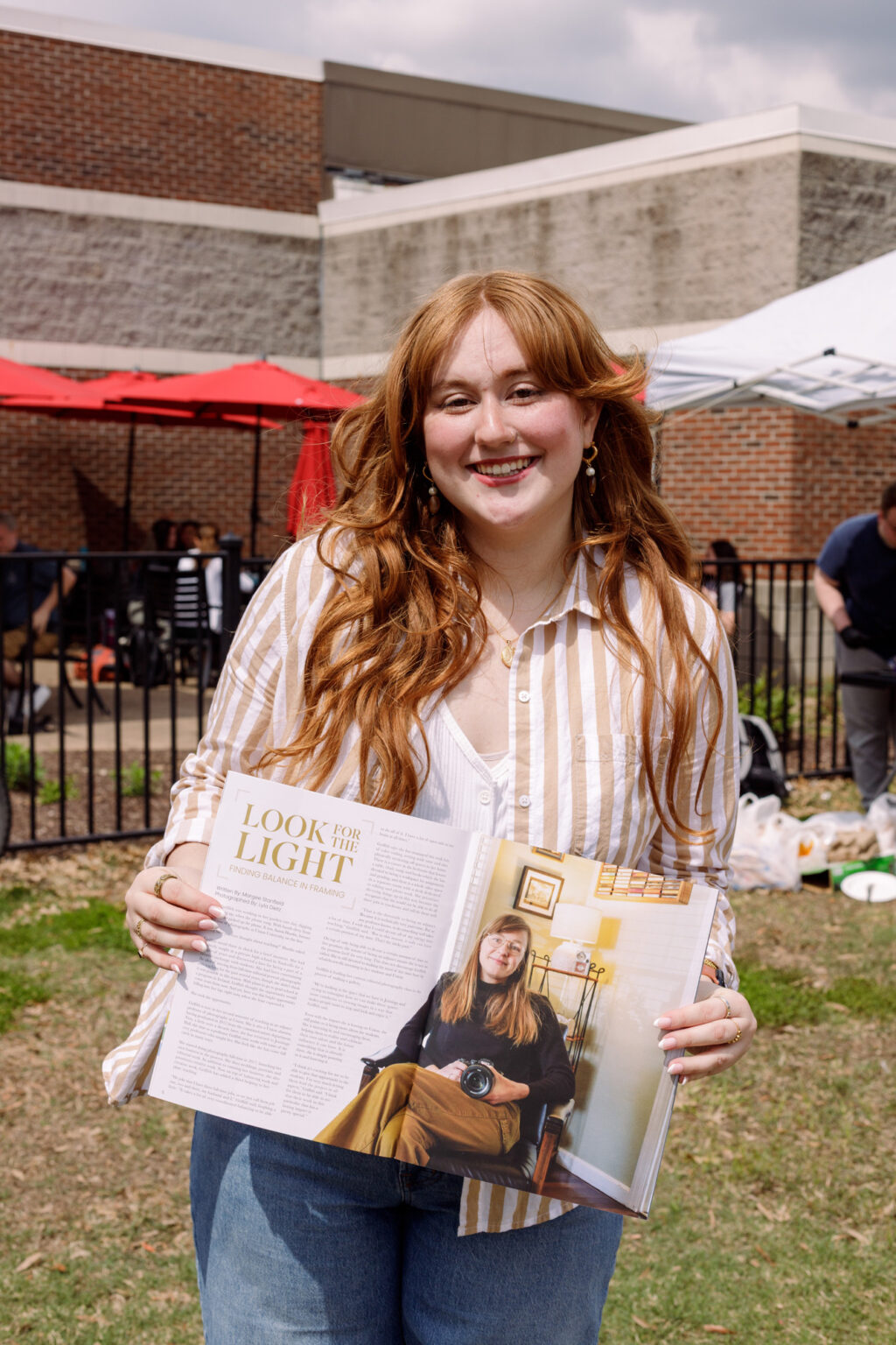 A person with long red hair stands outside, smiling and holding open a magazine that features a photo and article about another individual—a scene that highlights the impact of photojournalism. The background includes a building, tents, and people, suggesting an event where digital media communications play a key role in sharing stories.