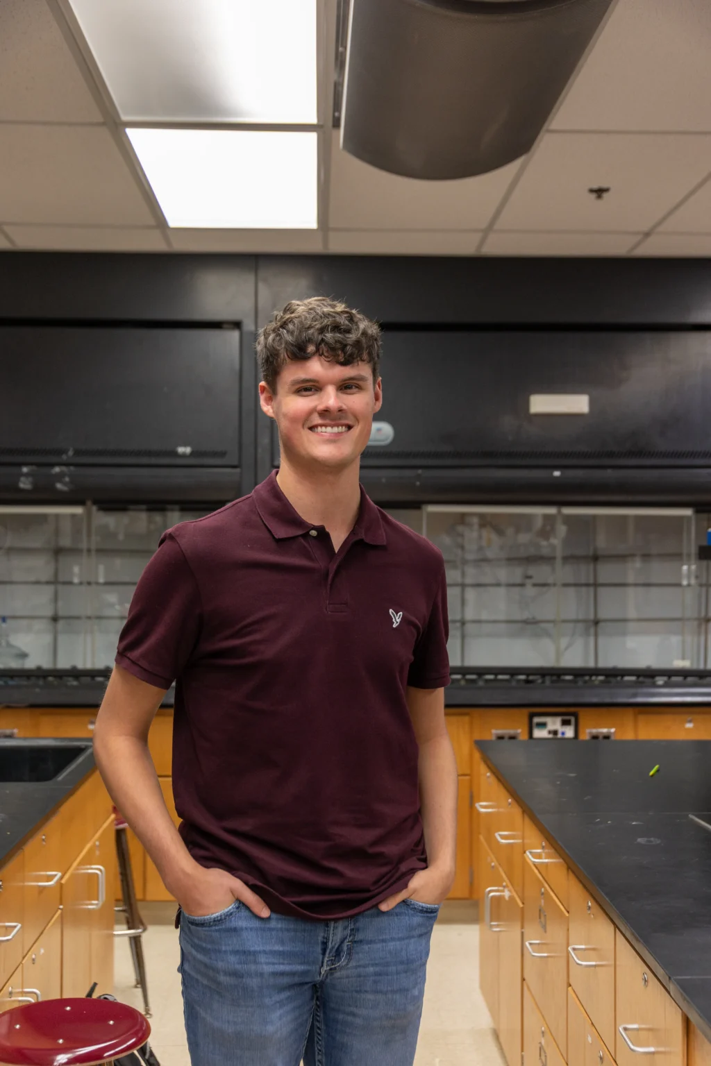 A person with short brown hair, wearing a maroon polo shirt and blue jeans, stands in a science classroom with wooden cabinets, black countertops, and laboratory equipment in the background.