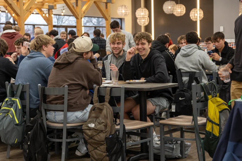 A group of young people sit around tables in a busy cafeteria, eating and talking. Some are smiling and laughing. Backpacks are on the floor and chairs, and the room has wooden beams and spherical hanging lights.