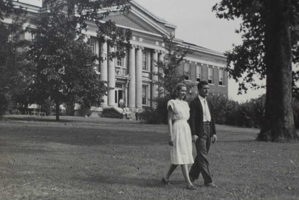 A woman in a light dress and a man in dark pants and a jacket walk hand-in-hand on a grassy lawn before a columned building, evoking the rich history of Union University. The photo is black and white, with trees framing the scene.