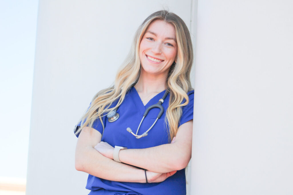 A woman in blue medical scrubs with a stethoscope around her neck, stands with arms crossed and smiles at the camera, leaning against a white wall.