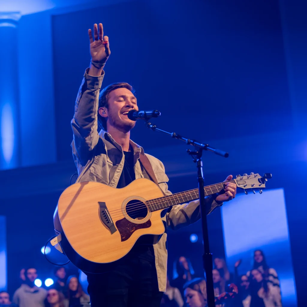 A man stands on stage playing an acoustic guitar and singing into a microphone, one hand raised. Blue stage lights illuminate the scene, capturing the vibrant energy of campus life as a group of people enjoy the performance in the background.