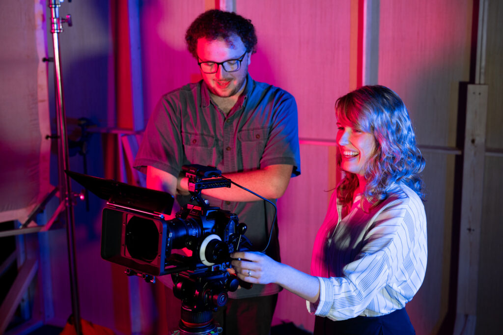 Two people stand behind a professional video camera in a studio with pink and blue lighting, capturing the essence of digital media communications. One person adjusts the camera while the other looks at the screen and smiles. Wooden panels and equipment are visible in the background.