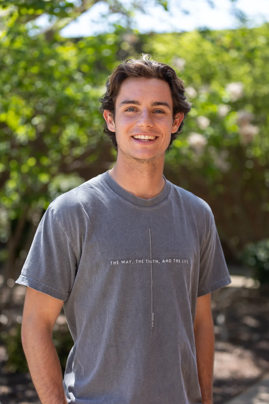 A young man with wavy brown hair smiles at the camera outdoors, wearing a gray T-shirt that reads THE WAY, THE TRUTH, AND THE LIFE. Green trees and sunlight are visible in the blurred background.