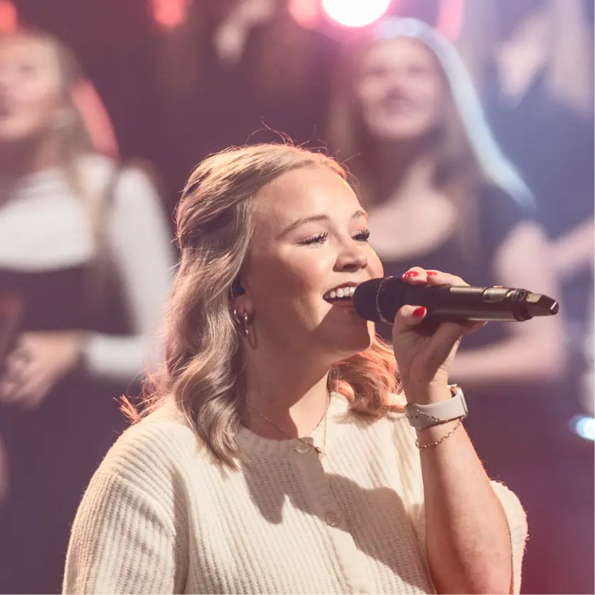 A woman with blonde hair sings into a microphone on stage. She is wearing a cream sweater and a white smartwatch. Several people are standing and blurred in the background, with stage lights visible.