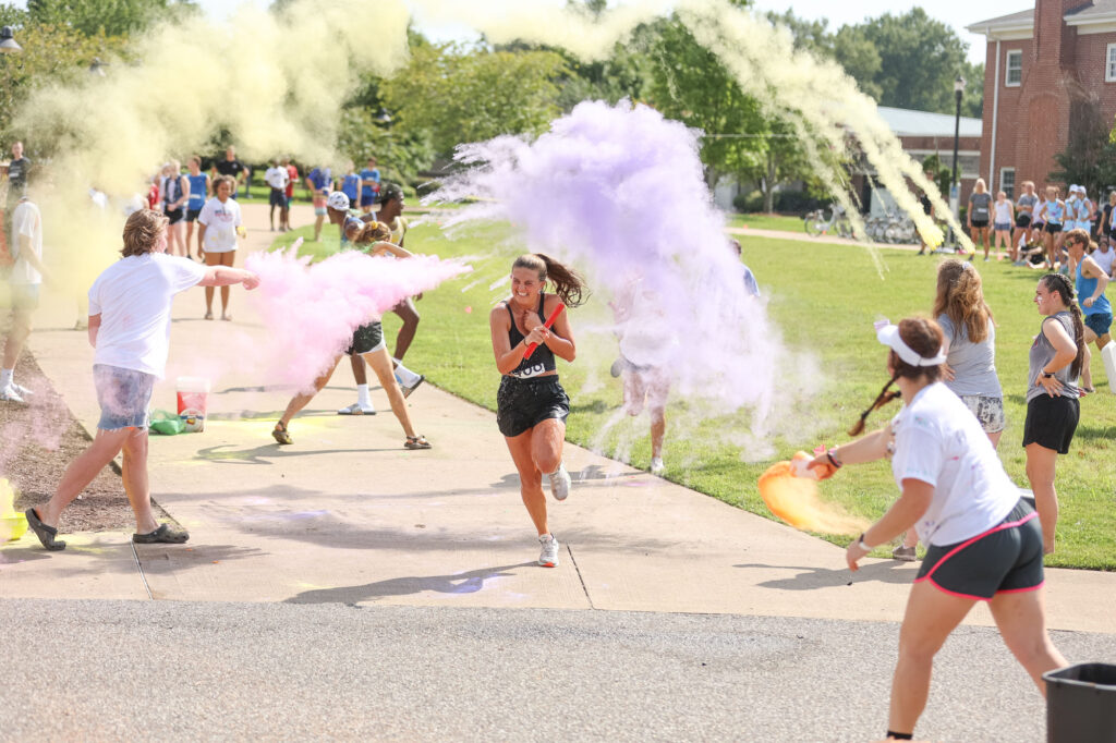 A group of people are throwing colored powder at participants in an outdoor event. One person is running through clouds of yellow and purple powder, while others stand nearby on a sunny day.