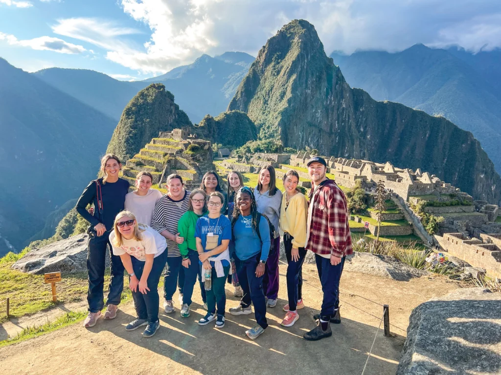 A group of people poses for a photo in front of the ancient ruins of Machu Picchu in Peru, with green mountains and blue sky in the background.