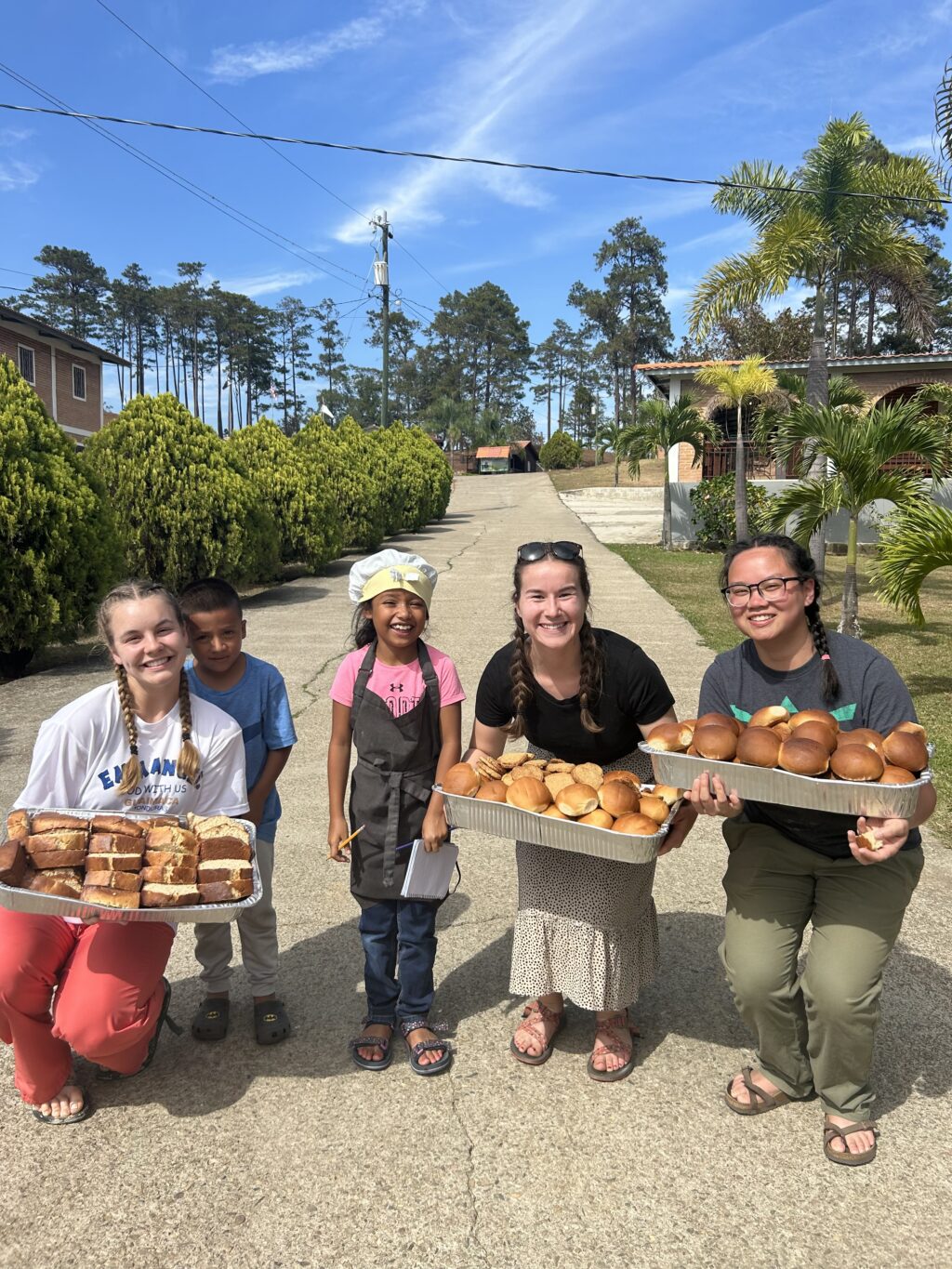 Four adults and one child stand outdoors on a sunny day, smiling and holding trays filled with bread and sandwiches—sharing food as part of their Christian Ministry. They are on a paved path surrounded by greenery and buildings in the background.