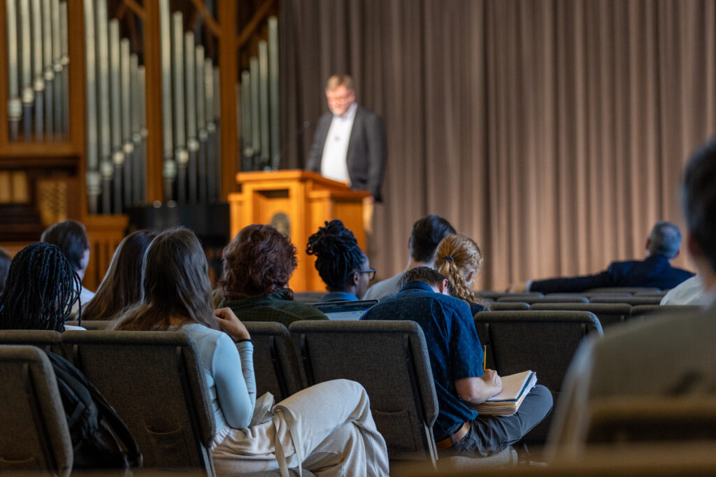 A group of people sit in rows facing a speaker standing at a podium on a stage. Some attendees are taking notes. An organ with pipes and closed curtains are visible in the background.