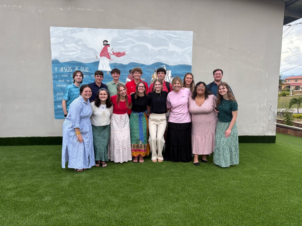 A group of fifteen people stands in two rows on green grass, smiling at the camera. Behind them is a mural showing a seaside scene and Spanish text. Most are dressed in colorful, modest clothing, reflecting their shared purpose in Christian ministry.